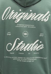 Πράσινο φούτερ με λευκό κείμενο που γράφει Originals Studio, Everyday Essentials Creative Dept. Holmen και φράσεις για άνεση και καθημερινή χρήση.