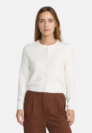 Frau mit langen, hellbraunen Haaren, die eine weiße, geknöpfte Strickjacke mit Lochmuster und braune hoch taillierte Hose vor weißem Hintergrund trägt.