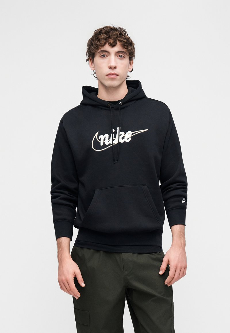 Black hoodie with a front pocket and a white Nike logo. Features a drawstring hood and ribbed cuffs. Paired with green cargo pants.