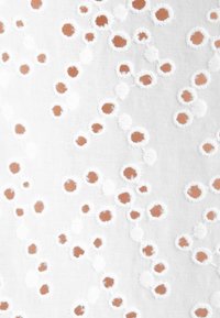 Tissu blanc avec de petites découpes circulaires brunes irrégulières bordées de points de couture blancs brodés, éparpillées sur la surface.