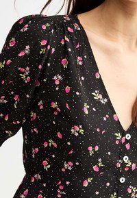 Vestido de tela negra con patrones florales en rosa y pequeños puntos blancos. Escote en forma de V con hombros fruncidos y botones blancos en la parte delantera.