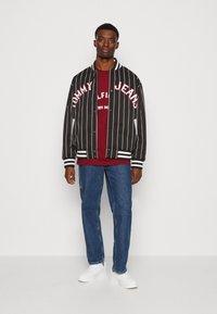 Mustvalge triibuline bomberjakk koos "TOMMY JEANS" logo ja burgundi värvi särk ning sinised teksad. Valged spordijalatsid täiustavad kogu välimust.