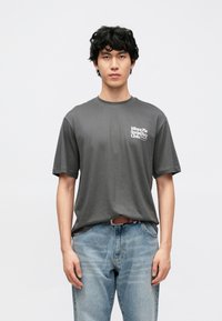 Sivé bavlnené tričko s krátkymi rukávmi, s bielym grafickým logom na ľavej strane hrudníka. Nosené s svetlomodrými rifľami.