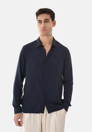 Uomo in piedi che indossa una camicia blu navy scuro con bottoni e maniche lunghe e pantaloni beige chiaro, con le mani rilassate lungo i fianchi.