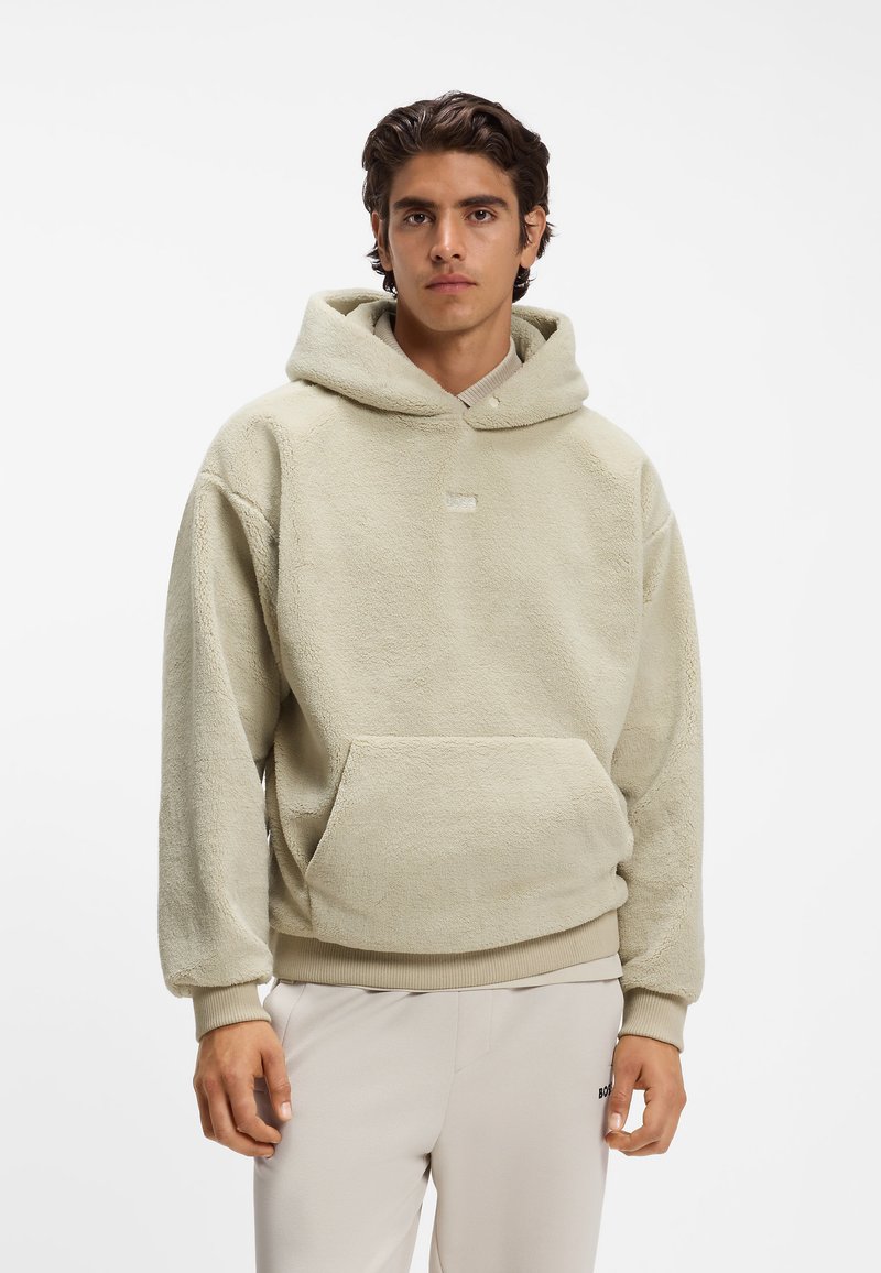 Νέος άνδρας που φοράει μπεζ φλις hoodie και ανοιχτόχρωμο παντελόνι στέκεται μπροστά σε απλό λευκό φόντο.