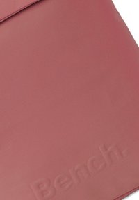 Sac rouge texturé avec une surface lisse, présentant un design de logo en relief subtil. Forme rectangulaire avec un système de fermeture à rabat.