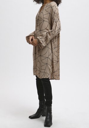Robe longue beige avec un motif abstrait noir, décolleté en V, manches surdimensionnées, fentes latérales, associée à un pantalon skinny noir et des bottines.