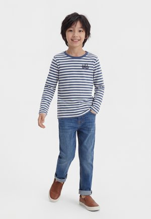 Smilende dreng iført en blå og hvidstribet langærmet skjorte med traktorprint, blå jeans og brune slip-on sko.