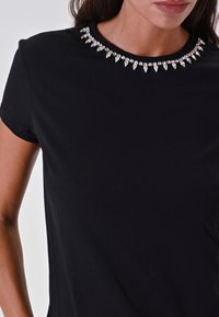 Camicia nera a maniche corte con un colletto decorativo caratterizzato da accenti simili a cristalli in un motivo a punte, realizzata in un tessuto liscio e elastico.