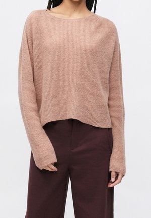 Femme portant un pull léger, côtelé, à manches longues couleur brun clair avec un pantalon large taille haute brun foncé sur fond blanc.