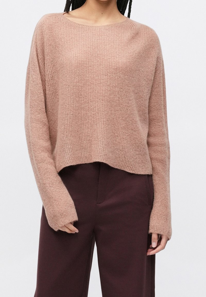 Femme portant un pull léger, côtelé, à manches longues couleur brun clair avec un pantalon large taille haute brun foncé sur fond blanc.