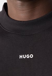 Gros plan d'un sweat-shirt noir à col rond avec le texte blanc « HUGO » imprimé sur la poitrine, porté par une personne à la peau foncée.