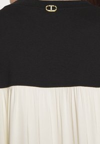 Sort topdel med rund hals, der fører ned til en plisseret cremefarvet stof nedenunder. Har et guld hardware-detalje i ryggen ved halsen.