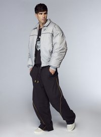 Giacca a vento argentata con collo alto, felpa nera con grafica e pantaloni neri larghi con piping laterale giallo e sneakers bianche.