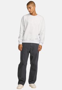 Light grey sweatshirt with crew neck and raglan sleeves, paired with dark grey pants. Model stands with hands in pockets and wears white sneakers.