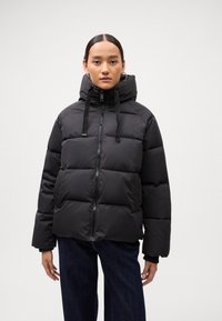 Black puffer jacket with a high collar, front zipper, and drawstring. Features quilted sections and a relaxed fit. Worn over dark denim.