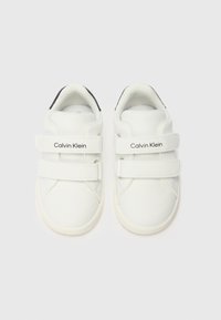 Baskets blanches en cuir avec double bande Velcro, bout rond, semelle légère et accent noir sur le talon. Texte "Calvin Klein" sur les bandes.