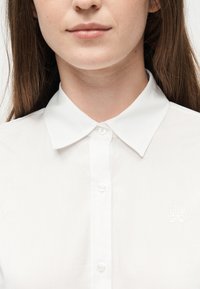 Γυναίκα που φοράει λευκό πουκάμισο με γιακά και κουμπιά, με το ανάγλυφο λογότυπο "HUGO" στο στήθος, εμφανιζόμενη από το πηγούνι έως το άνω μέρος του κορμού.