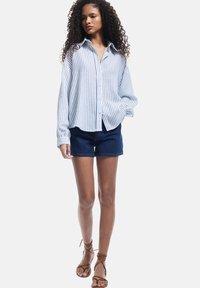 Stribet skjorte med knapper i lyseblå og hvid, parret med mørke denimshorts. Modellen har brune sandaler med remme på. Krøllet, løst hår.