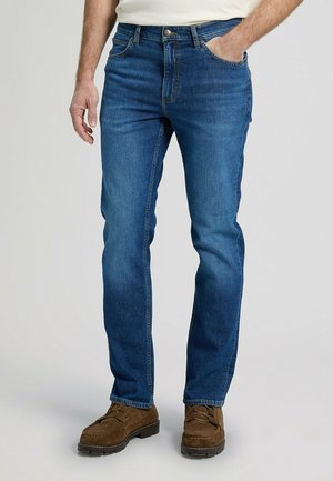 Mann trägt mittelblaue Jeans mit geradem Bein, weißes Hemd eingesteckt und braune Wildleder-Schnürschuhe, steht mit einer Hand in der Tasche.