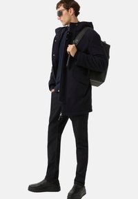 Mężczyzna w ciemnej, casualowej kurtce, w okularach przeciwsłonecznych, z czarnym plecakiem, ciemnymi dżinsami i czarnymi butami, stojący bokiem z jedną ręką w kieszeni.