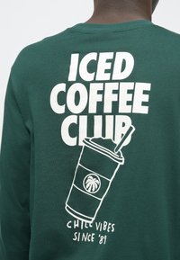 Zeleno majica dugih rukava s bijelim grafičkim natpisom "ICED COFFEE CLUB" i ilustracijom šalice koja prikazuje palmu i slamku.