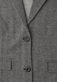 Gros plan d'un manteau gris à motif chevrons avec deux boutons noirs et deux petites poches avant visibles.