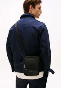 Muž v námořnické bundě a bílém tričku nese černou crossbody tašku Tommy Hilfiger, stojí bokem na bílém pozadí.