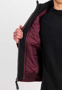 Sort jakke med lynlås fortil, med et maroon-farvet for og ribbede manchetter. Jakken åbner for at afsløre en quiltet indre tekstur.