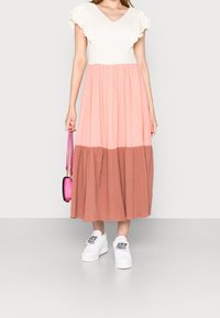 Abito maxi con maniche corte a balze, caratterizzato da una parte superiore bianca, una sezione centrale rosa e una gonna marrone. Abbinato a sneakers bianche e una borsa rosa.