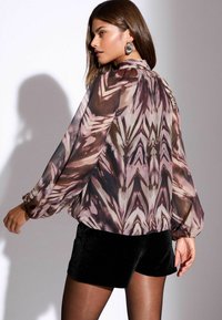 Lange, musterige blouse lavendli ja pruuni tooni, millel on kogutud varrukad ja kõrge kaelus, sobitatud mustade lühikeste pükstega.