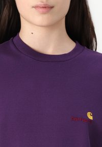 Close-up van een persoon in een paarse Carhartt t-shirt, waarbij de halslijn en het geborduurde logo op de borst zichtbaar zijn.