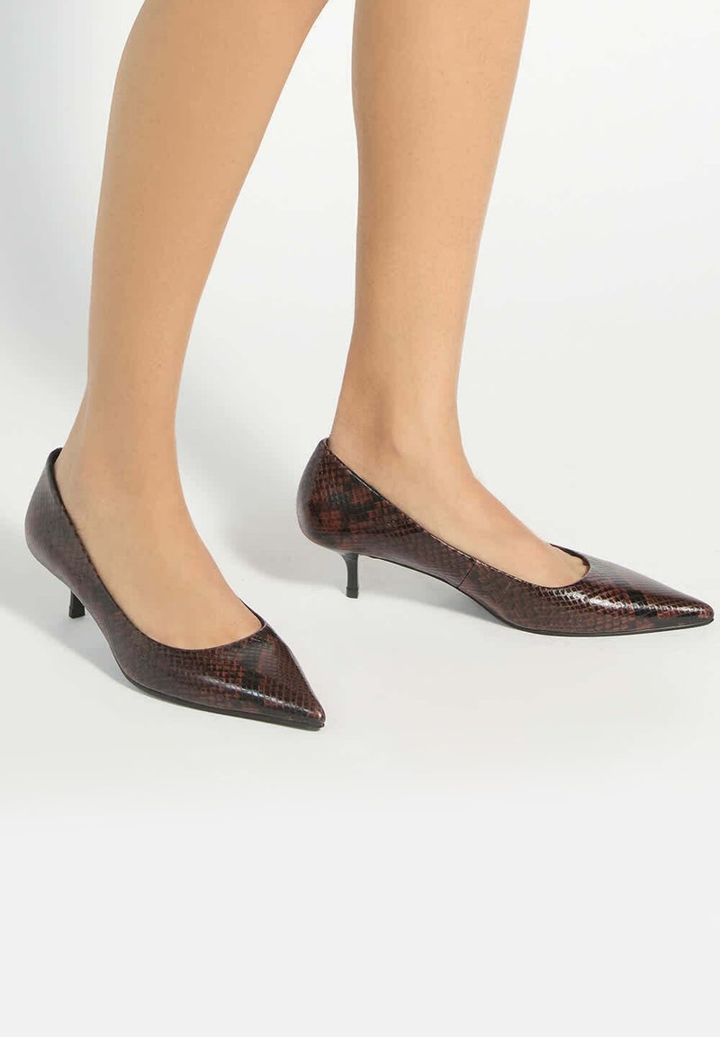 Jambes fines portant des chaussures à talons kitten à motif peau de serpent marron foncé à bout pointu sur un fond clair.
