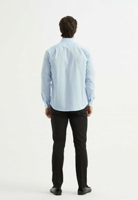 Chemise bleu clair à manches longues avec col boutonné et ourlet incurvé, associée à un pantalon noir ajusté et des chaussures noires, vue de dos.