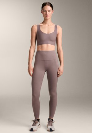 Woman standing wearing mauve sports bra and high-waisted leggings with white athletic shoes against neutral background.