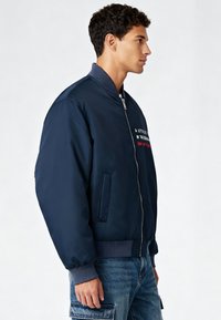 Veste bomber bleu marine avec une fermeture éclair à l'avant, des poignets côtelés et un texte brodé sur la poitrine. Constituée d'un matériau lisse et brillant.