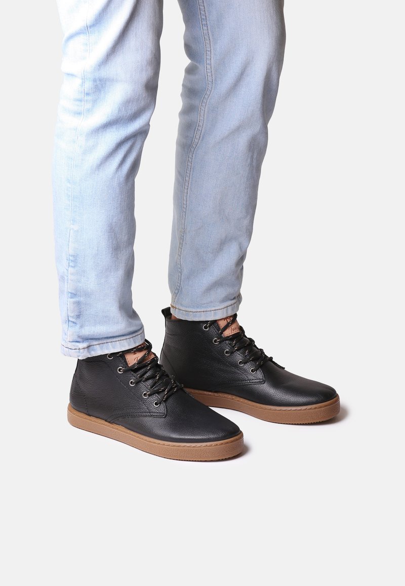Botas de tobillo de cuero negro con puntera redonda y cuello elevado, con suela de goma marrón y cordones con ojetes. Llevadas con jeans de color azul claro.