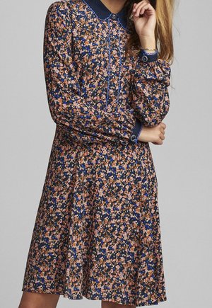 Femme portant une robe à manches longues à motifs floraux avec col et poignets bleu marine, comportant des couleurs roses, bleues et noires, debout les bras croisés.