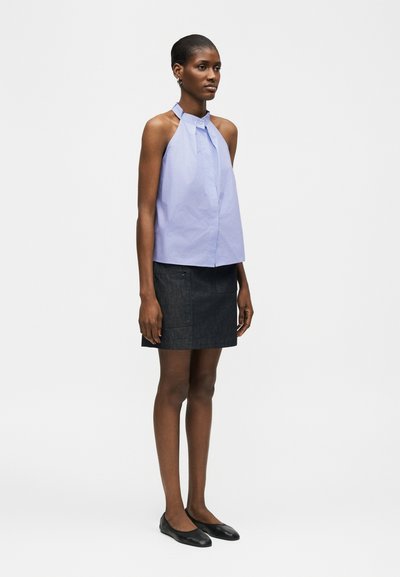 Femme en haut sans manches bleu clair à col halter et mini-jupe noire, debout face à l'avant, les bras détendus le long du corps.
