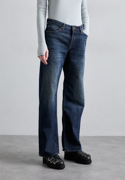 Jeans din denim de culoare albastră închisă, cu o croială largă, având cinci buzunare și o ușoară decolorare. Asortați cu pantofi negri și un top gri deschis.