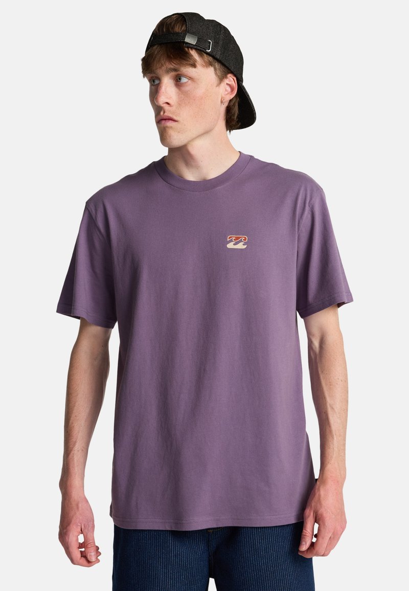Jeune homme portant un t-shirt violet avec un petit logo de vague sur la poitrine et une casquette noire portée à l'envers, regardant vers la gauche.