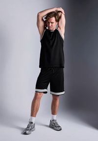 Μαύρο αθλητικό τοπ χωρίς μανίκια και σορτς με λευκές ρίγες. Το υλικό φαίνεται ελαφρύ· η ενδυμασία είναι σχεδιασμένη για άνεση και ελευθερία κινήσεων. Γκρι αθλητικά παπούτσια.