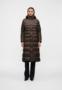 Lang brun pufferjakke med høj krave og knaplukning, med en quiltet design og hætte, parret med mørke jeans og spidse hæle.