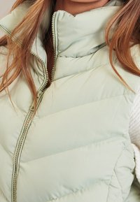 Gilet imbottito verde menta con collo alto, chiusura frontale con zip e motivo trapuntato orizzontale; tessuto liscio e design leggero.