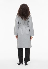 Kvinne med krøllete hår iført en belted grå frakk, svarte bukser og svarte høye støvler, stående med ryggen mot kameraet mot en hvit bakgrunn.