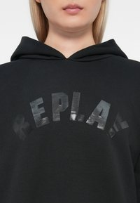 Černá mikina s kapucí, která má jemnou texturu a výrazné lesklé tiskové logo s nápisem "REPLAY" na hrudi v zakřiveném designu.