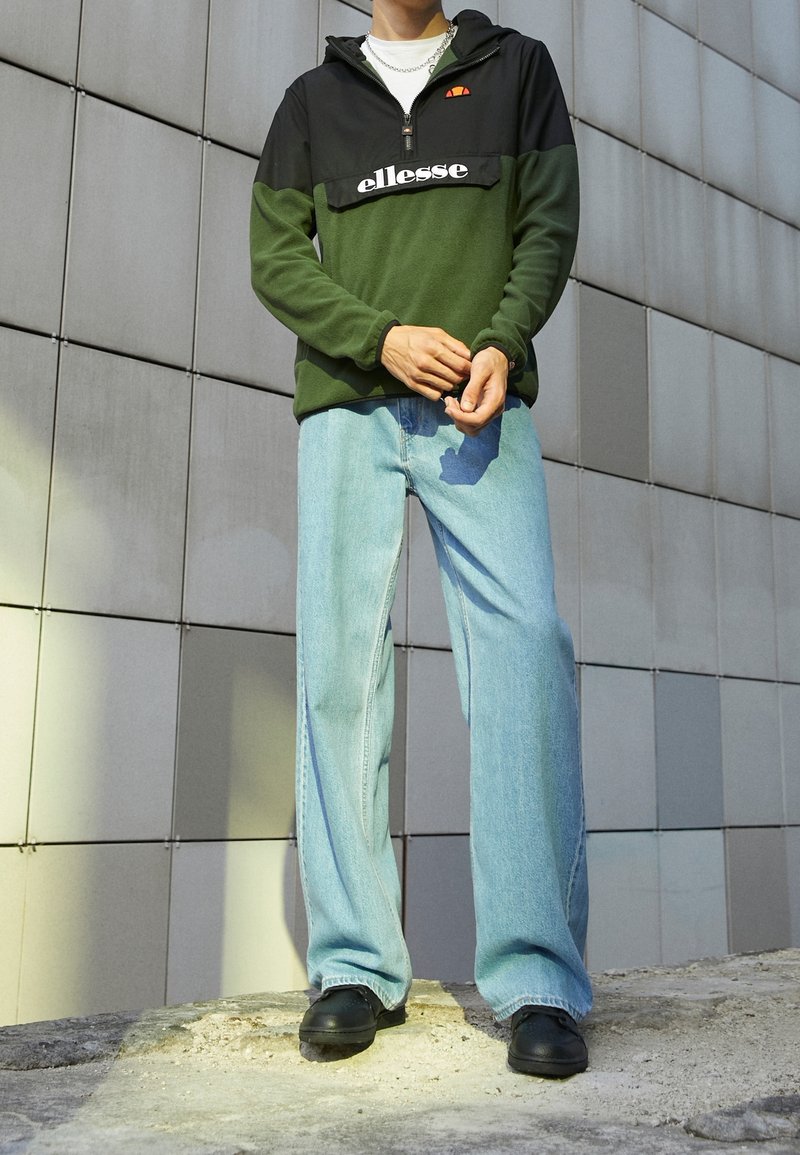 Zielono-czarna bluza polarowa z suwakiem przy szyi i białym napisem „ellesse”. Jasnoniebieskie szerokie dżinsy o luźnych nogawkach i czarne buty.