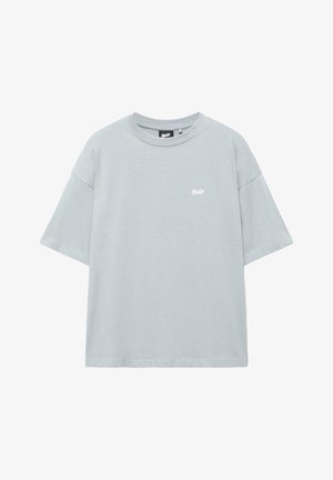 Lichtgrijs oversized T-shirt met korte mouwen, ronde hals en klein wit logo op de linkerborst, tegen een witte achtergrond.