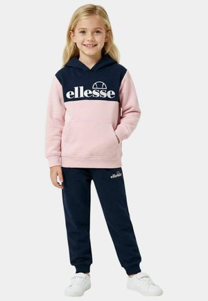 Jeune fille souriante portant un sweat à capuche Ellessee rose et bleu marine et un pantalon de survêtement bleu marine avec des baskets blanches, debout devant un fond uni.