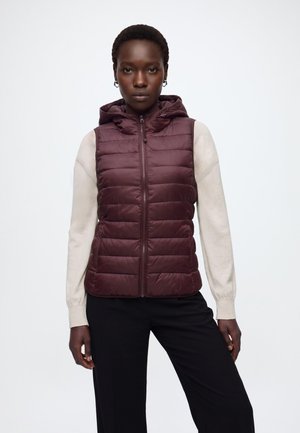 Femme portant une veste matelassée bordeaux sans manches avec capuche par-dessus une chemise crème à manches longues et un pantalon noir, debout devant un fond uni.